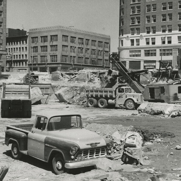 Black-and-white photo of a city construction site with rubble, a vintage pickup truck in the foreground, dump trucks, a crane, and tall office buildings in the background.