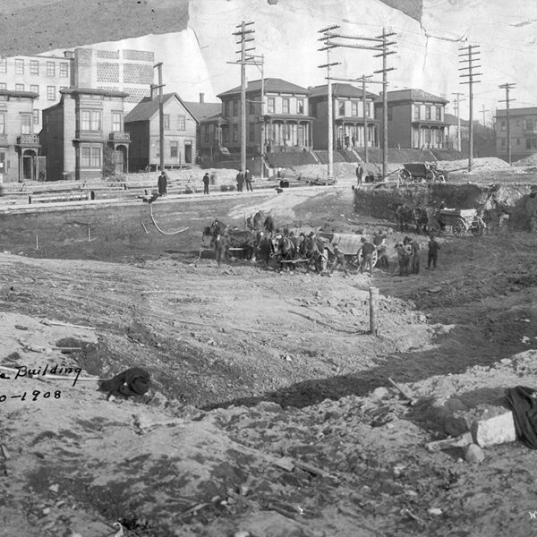 Historic black-and-white photo of a construction site with workers, horses, and wagons in front of early 1900s homes. Handwritten note reads: Site White Building Jan. 30 - 1908. Utility poles and dirt foreground visible.