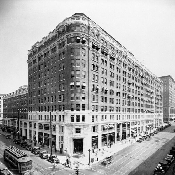 A black-and-white photo of a large, multi-story brick building on a busy downtown corner with early 20th-century cars, streetcars, and people on the streets and sidewalks.