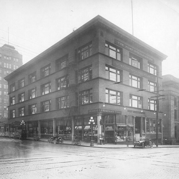 Black and white photo of a four-story brick building on a street corner, with early 1900s cars parked outside. Signs read The Post-Intelligencer. Other historic buildings and street lamps are visible nearby.