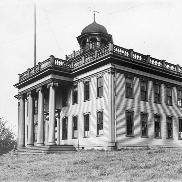 Historic black-and-white photo of a large, two-story building with tall columns, a central cupola, and a balustraded roof, set on a grassy hill with a few trees nearby.