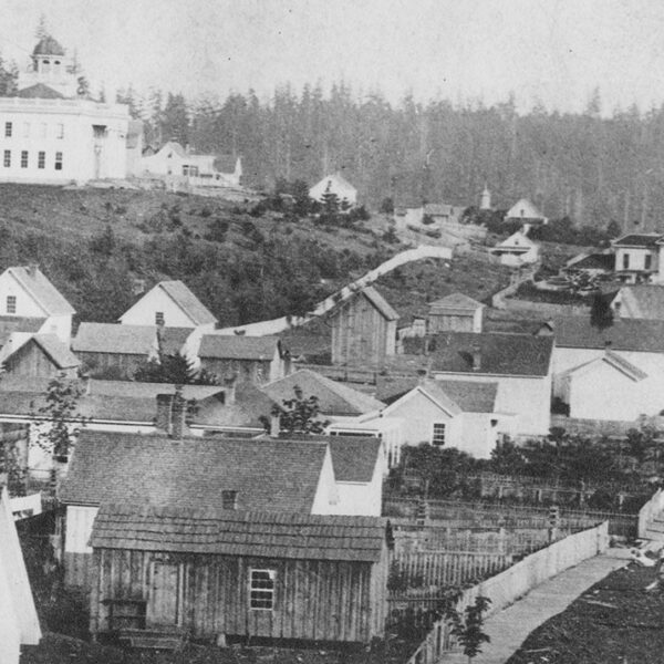 Black and white photo of a historic village with wooden houses, fences, and a hilltop building surrounded by trees and forest in the background. The scene suggests a rural settlement from the past.