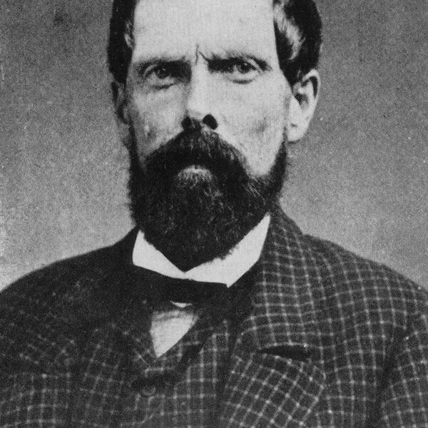 Black-and-white portrait of a man with a full beard and mustache, wearing a checkered suit, white shirt, and bowtie. He has short, dark hair and a serious expression, looking directly at the camera.