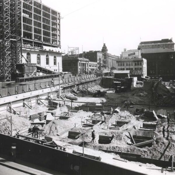 Black-and-white photo of a large urban construction site with deep excavations and wooden supports. Surrounding buildings and a crane are visible in the background. Several workers are present in the area.