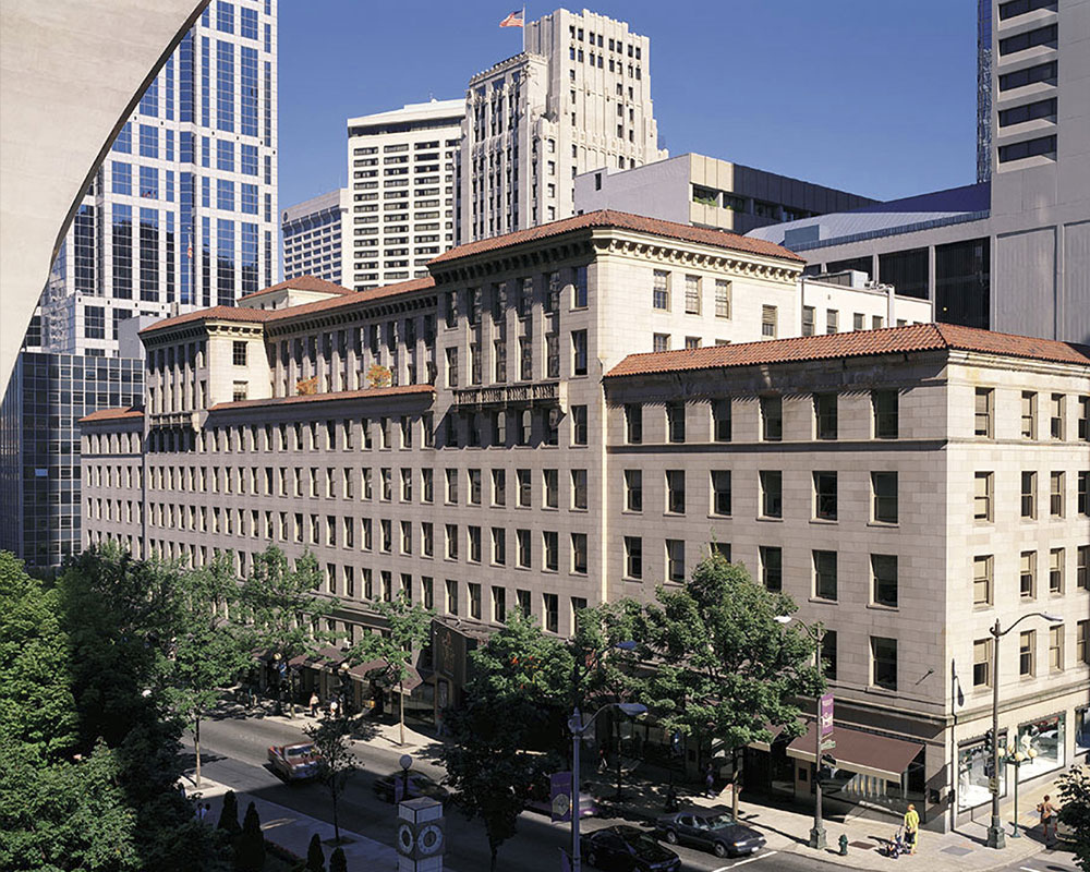A large, multi-story beige stone building with red-tiled roofs stands on a busy city street, surrounded by trees and tall modern skyscrapers under a clear blue sky.