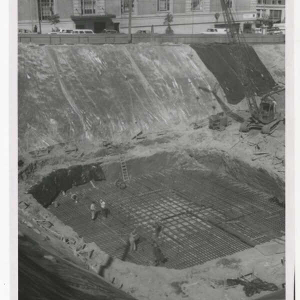 Black and white photo of a construction site with workers installing steel reinforcement bars in a large excavation pit; a crane and buildings are visible in the background.