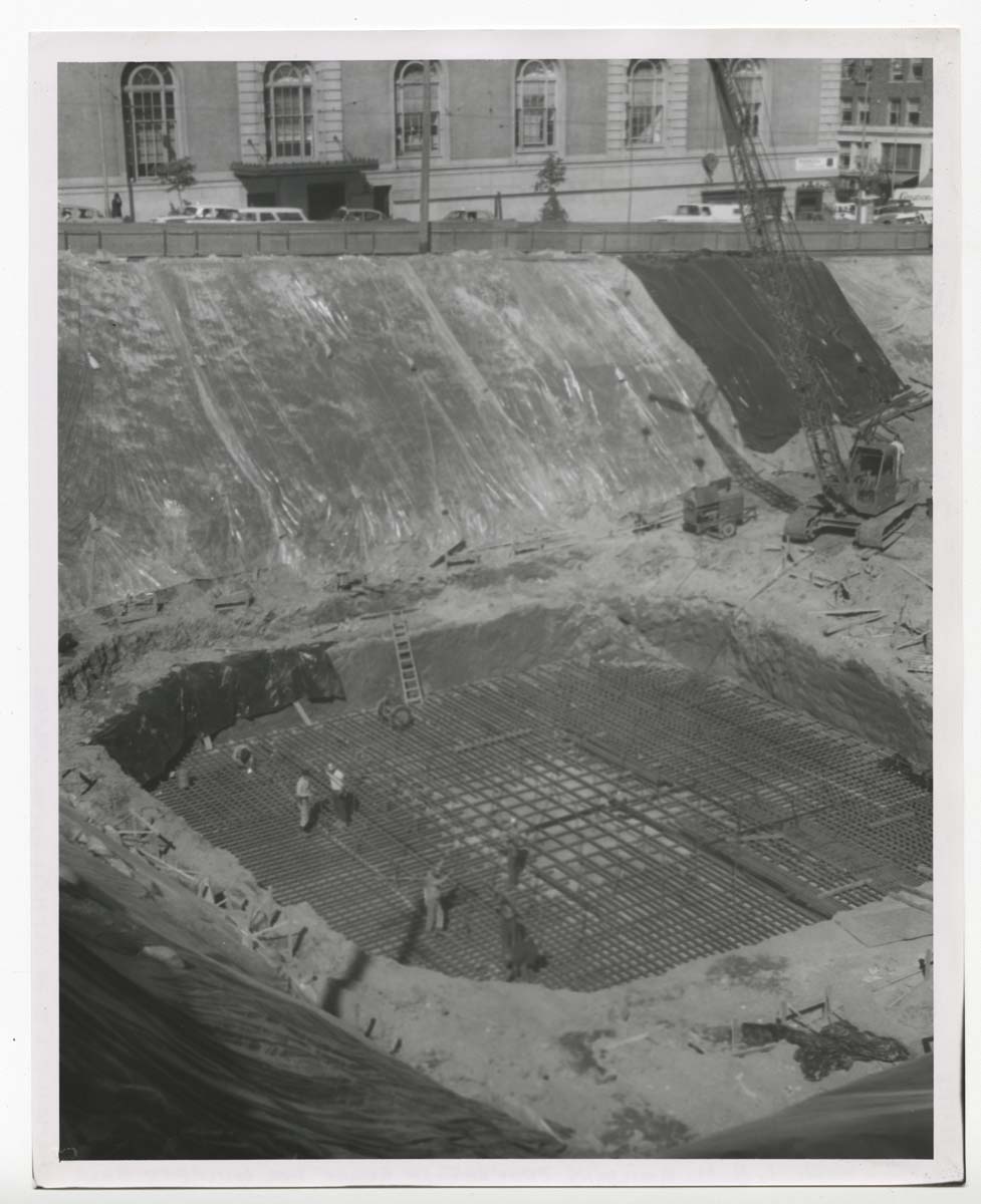 Black and white photo of a construction site with workers installing steel reinforcement bars in a large excavation pit; a crane and buildings are visible in the background.