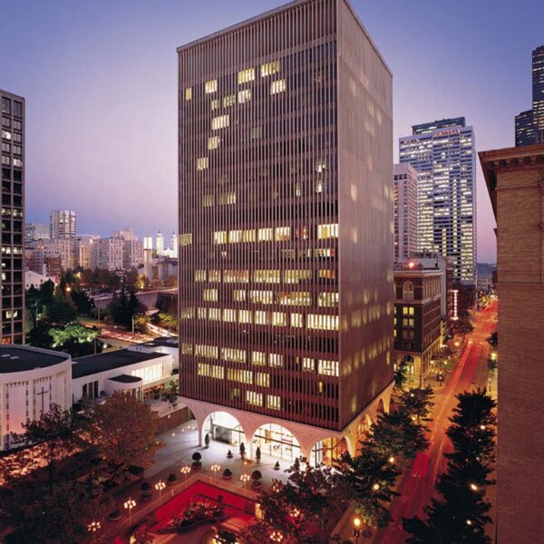 A tall office building with vertical lines and illuminated windows stands in a city at dusk, surrounded by other skyscrapers, trees, and a red-lit plaza with benches and a fountain.