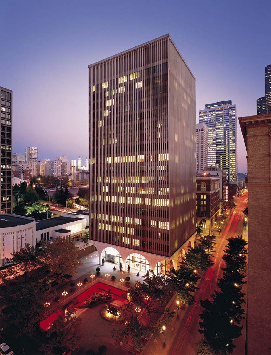 A tall office building with vertical lines and illuminated windows stands in a city at dusk, surrounded by other skyscrapers, trees, and a red-lit plaza with benches and a fountain.