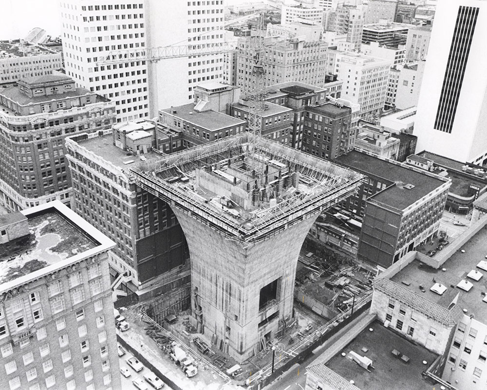A black-and-white aerial photo shows a tall building under construction in a city. The structure has a wide, flared top with scaffolding and cranes, surrounded by other mid- and high-rise buildings.