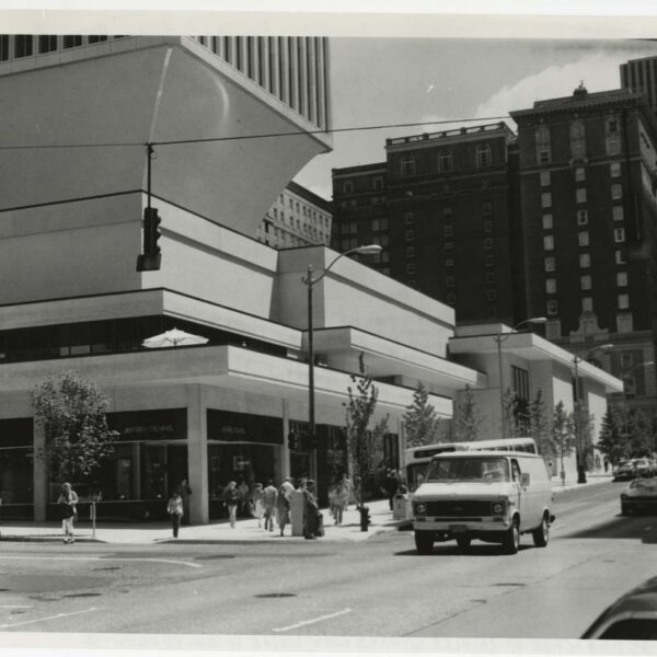 Black and white photo of a city street corner with modern, angular buildings, a group of people walking on the sidewalk, and a white van driving down the street. Tall older buildings appear in the background.