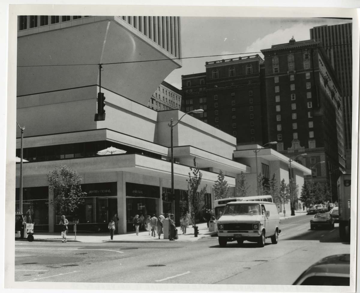 Black and white photo of a city street corner with modern, angular buildings, a group of people walking on the sidewalk, and a white van driving down the street. Tall older buildings appear in the background.