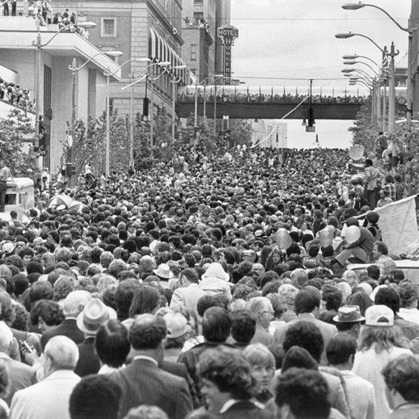 A large, dense crowd fills a city street during a public event or parade. People stand shoulder to shoulder, facing forward. Buildings, trees, and a bridge are visible in the background under a cloudy sky.