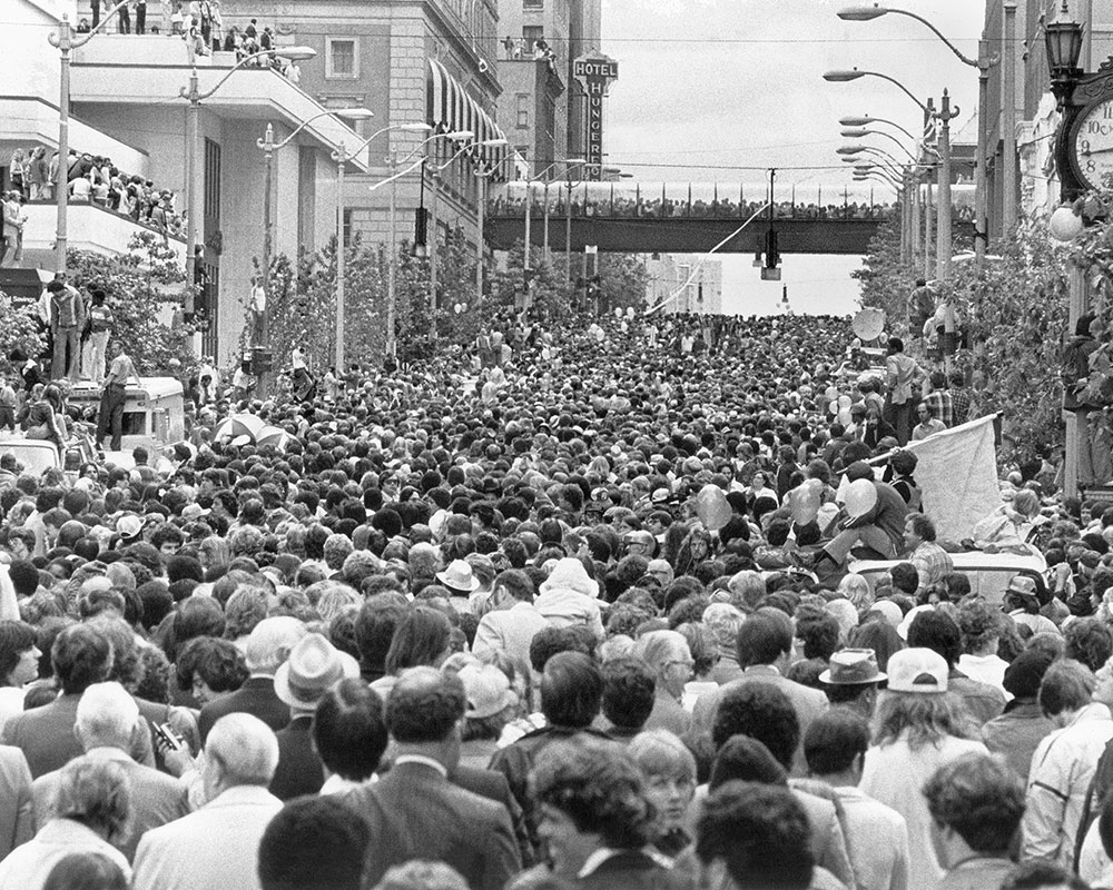 A large, dense crowd fills a city street during a public event or parade. People stand shoulder to shoulder, facing forward. Buildings, trees, and a bridge are visible in the background under a cloudy sky.