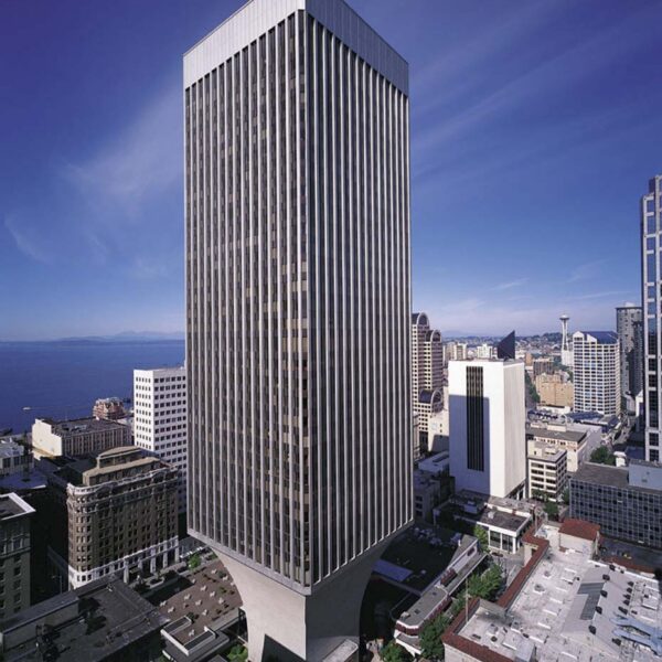 A tall, modern skyscraper with a unique flared base stands among other buildings in a cityscape under a clear blue sky. The structure is surrounded by urban streets, with distant water and more buildings in the background.