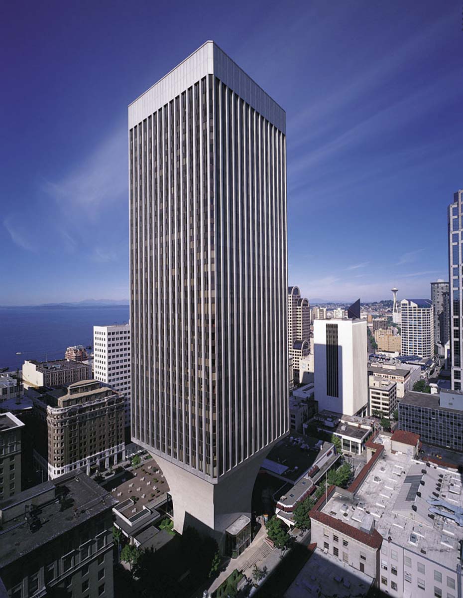 A tall, modern skyscraper with a unique flared base stands among other buildings in a cityscape under a clear blue sky. The structure is surrounded by urban streets, with distant water and more buildings in the background.