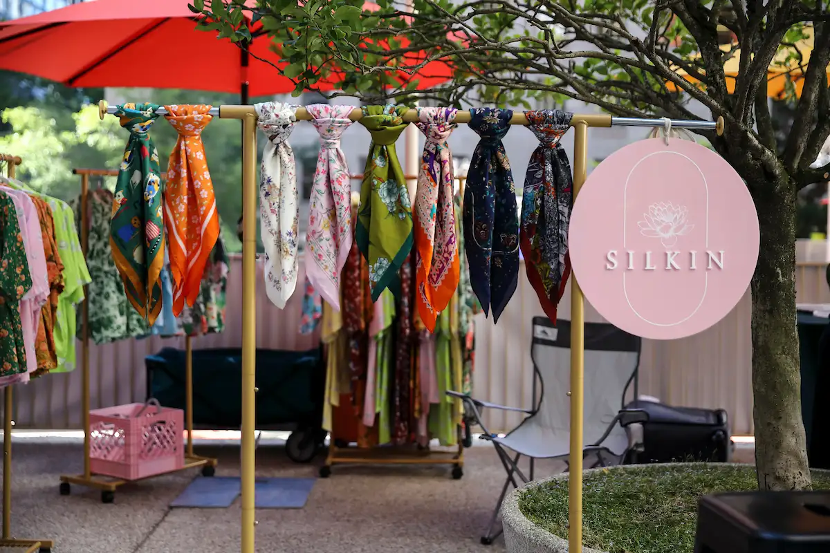 Colorful scarves hang on a rack under a tree at an outdoor market stall. A pink circular sign with SILKIN printed on it is displayed on the right side of the rack.