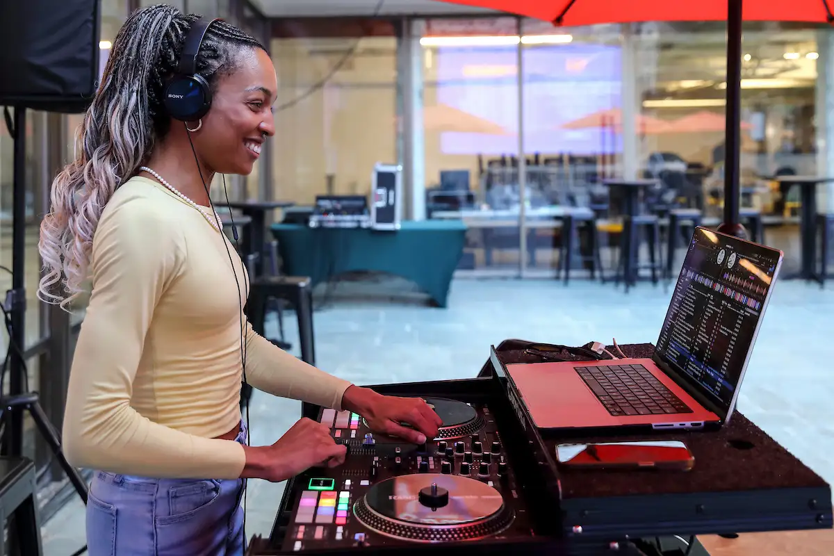 A smiling DJ wearing headphones mixes music on a controller and laptop at an outdoor event. Tables and chairs are visible through the windows behind her.