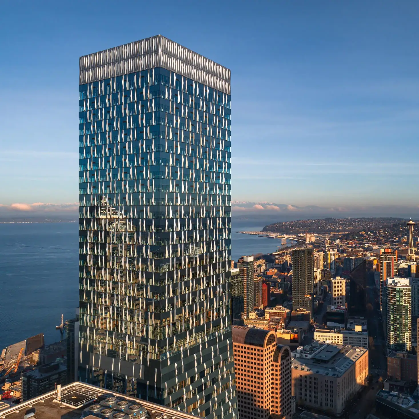 A tall, modern glass skyscraper reflects the sky and nearby buildings in a cityscape, with a coastline and the ocean visible in the background under clear blue skies.