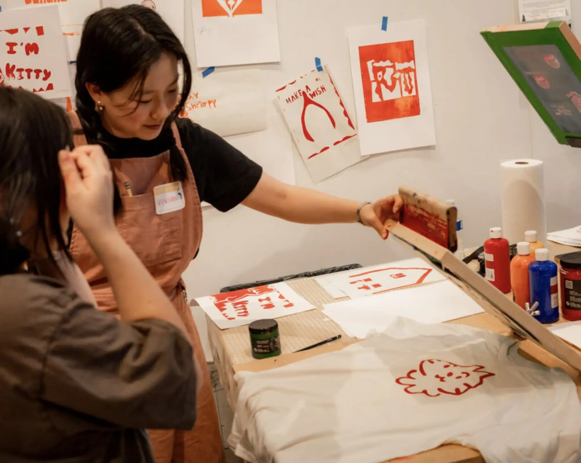 Two people screen print a red lion design onto a white T-shirt in an art studio. Finished prints and art supplies, including paint bottles and paper towels, are visible on the table and wall.