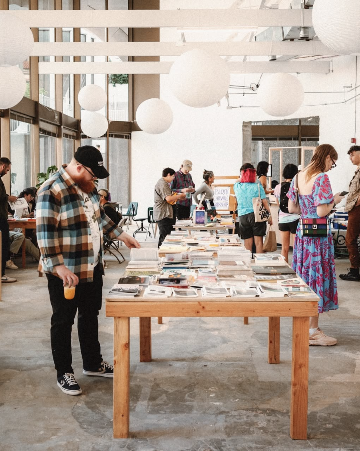 People browse books and magazines on tables in a bright, modern space with large windows and hanging white paper lanterns. Some stand and read, while others walk around or talk in small groups.