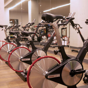 A row of stationary exercise bikes with red accents is lined up in the modern MT Fitness Center, reflected in a wall mirror above wooden flooring and bright lighting—available with an MT Fitness Center Membership.