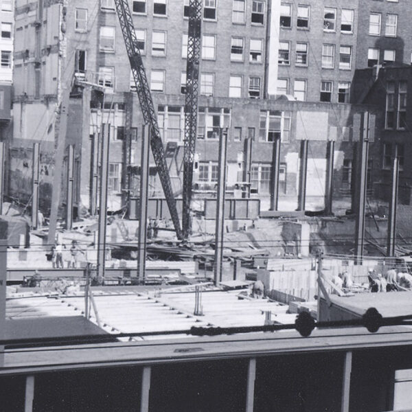 Black and white photo of a construction site in an urban area, with steel beams, cranes, and partially completed structures. Surrounding the site are tall office buildings and visible business signs.