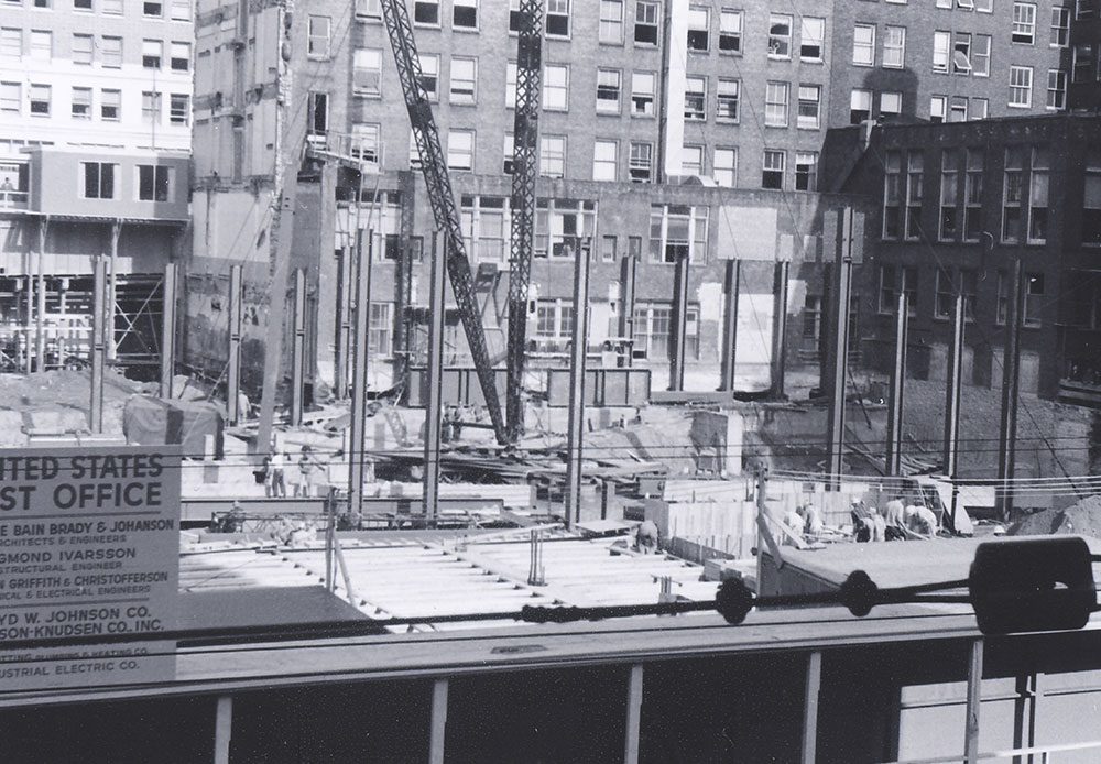 Black and white photo of a construction site in an urban area, with steel beams, cranes, and partially completed structures. Surrounding the site are tall office buildings and visible business signs.