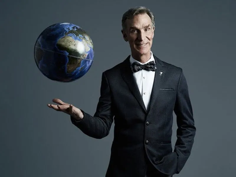 A man in a black suit and bow tie smiles while holding a floating globe with one hand against a plain gray background.