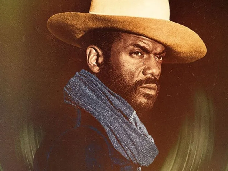 A man with a beard wearing a wide-brimmed tan hat and a blue scarf looks intensely to the side against a dark background.