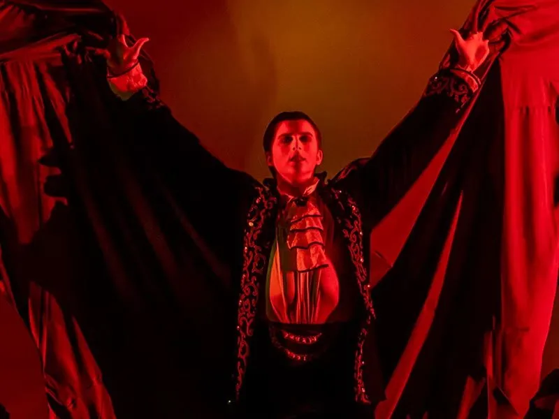 A person dressed as a vampire in dramatic lighting spreads a black cape wide, wearing a ruffled white shirt and ornate jacket, with a red background enhancing the theatrical, gothic atmosphere.