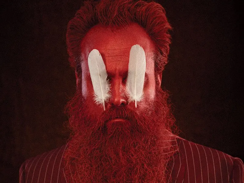 A man with a full red beard and hair wears a striped suit. Two large white feathers cover his eyes, set against a dark background.