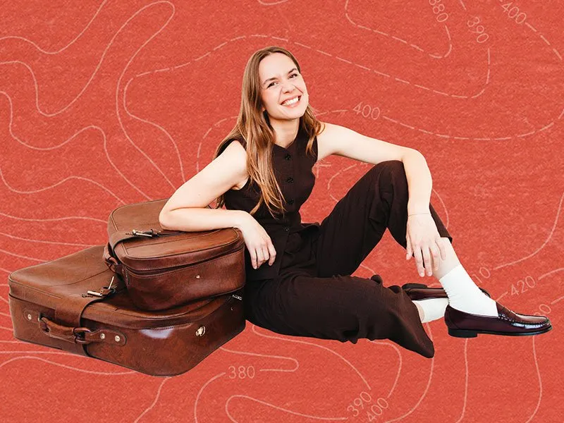 Smiling woman in a black jumpsuit sits on the floor, leaning on two stacked brown suitcases, against a textured red background with faint curvy white lines.