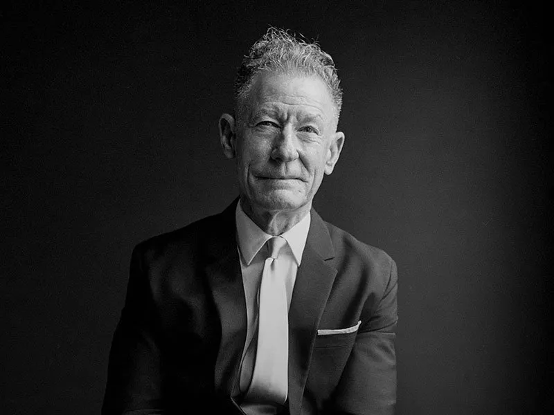 Black and white photo of an older man in a suit and tie, sitting against a dark background, facing the camera with a slight smile and curly hair.