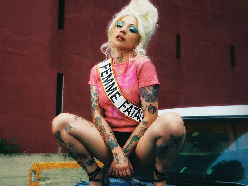 Tattooed person with platinum blonde hair and bold blue eyeshadow squats on a car, wearing a pink shirt and a sash reading FEMME FATALE, against a red wall background.