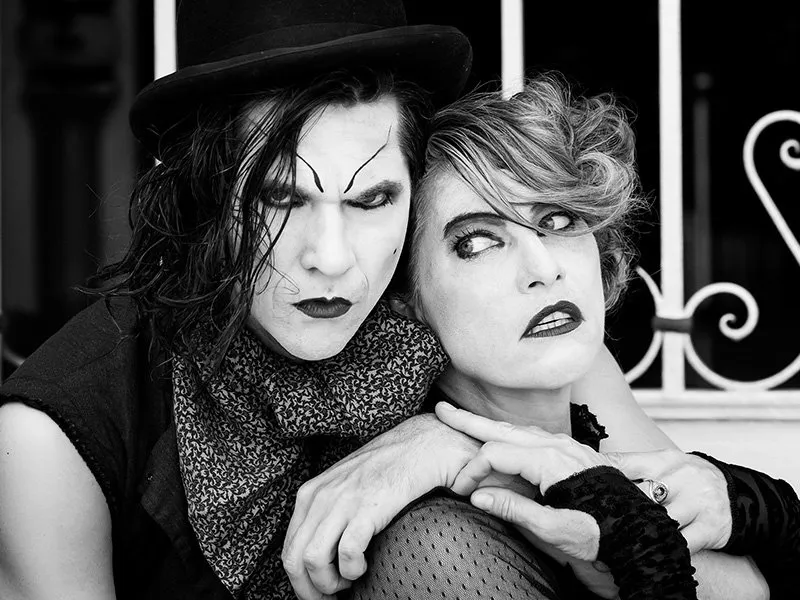 Two people in dramatic makeup and dark clothing pose closely together. One wears a top hat and scarf with sharp eyebrows, while the other has short, wavy hair and a mesh top. The background features white metal bars.