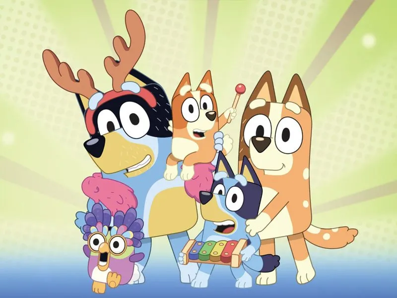 Four cartoon dogs and a bird, all wearing colorful costumes and smiling, pose together on a green background with rays and dots. One dog holds a xylophone, and the smallest rides on a larger dogs back, holding a drum mallet.